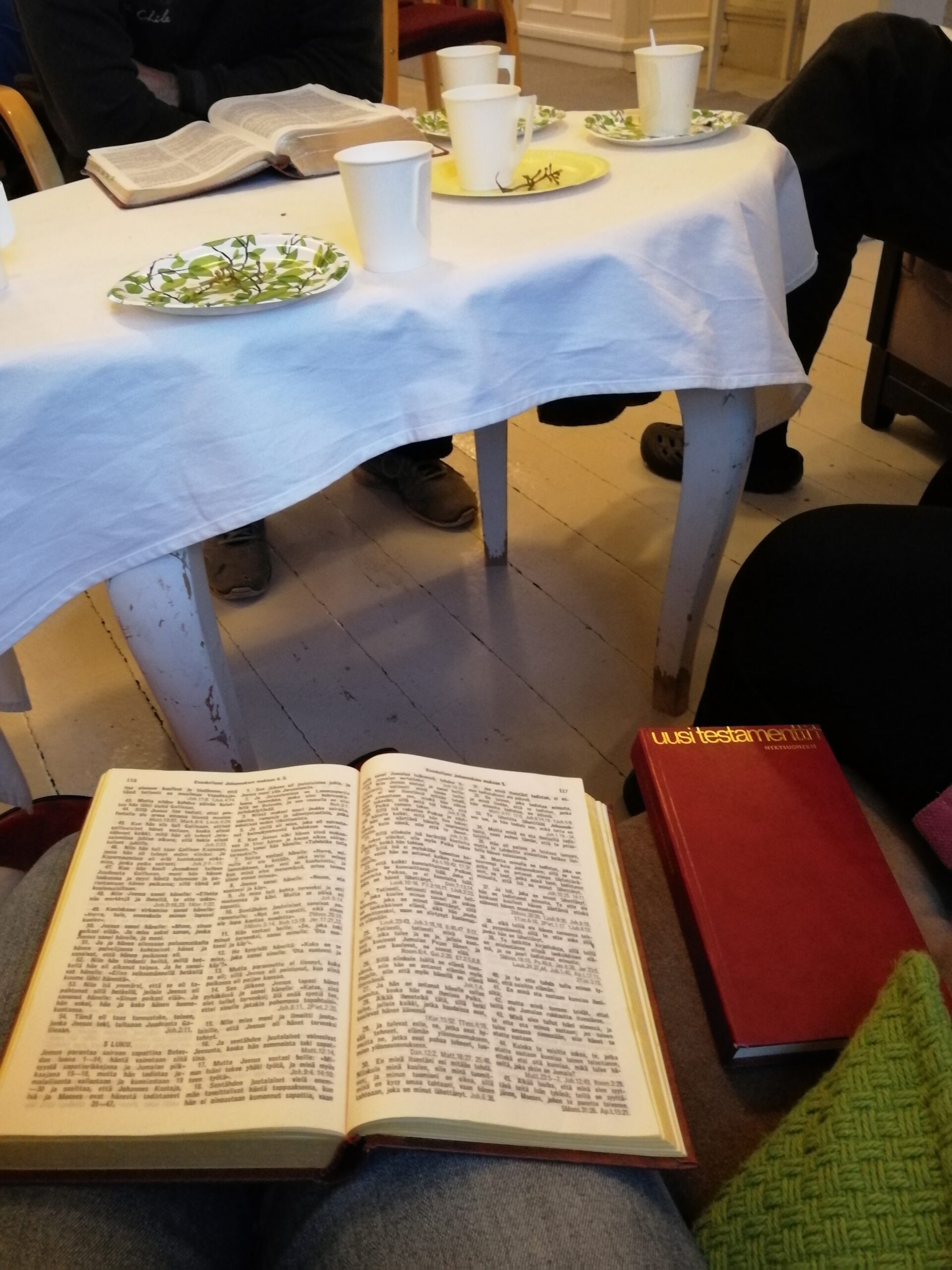 Raamattu ja tausttalla pöytä, jossa valkoinen pöytäliina.
