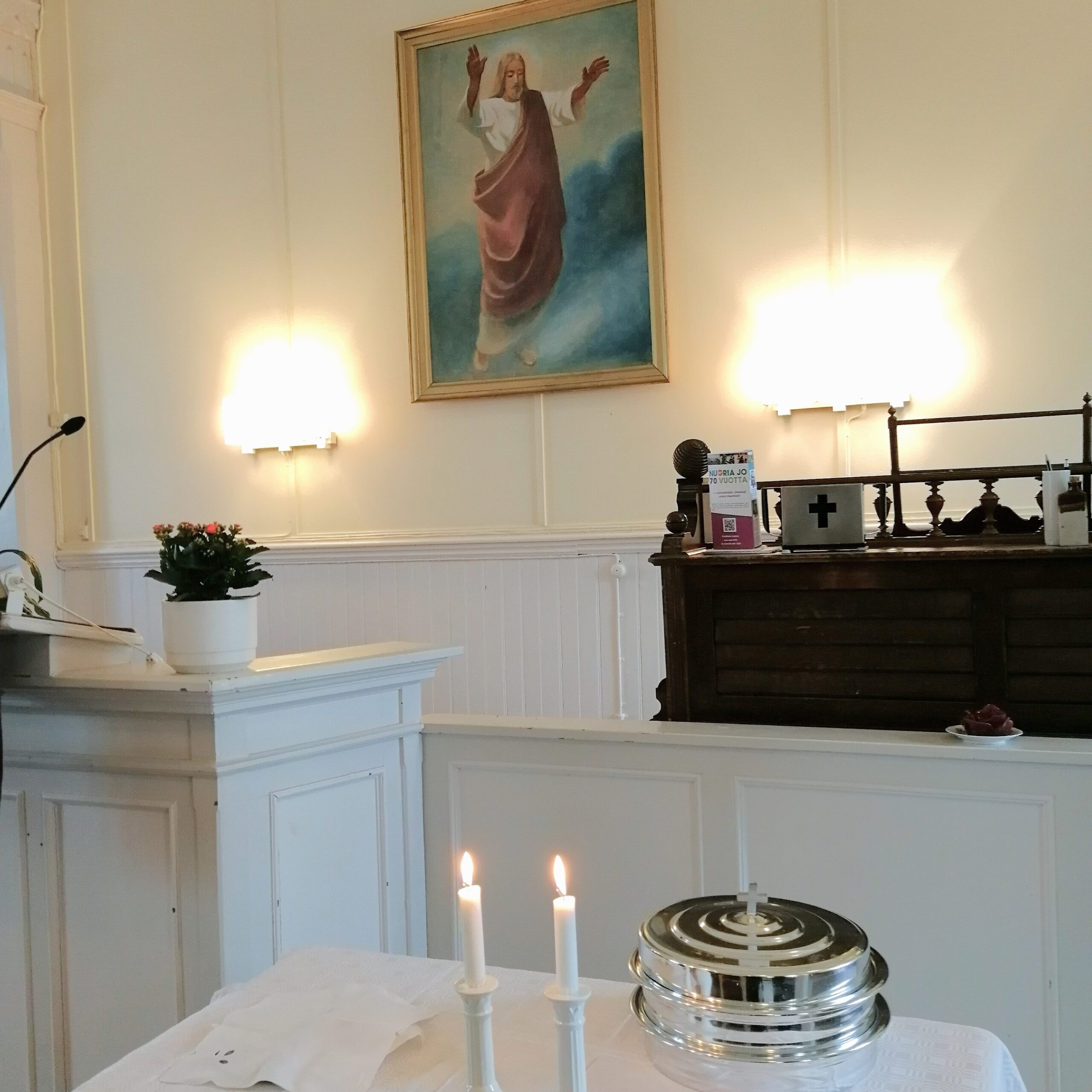Kirkkosalissa ehtoollispöytä, jossa kaksi kynttilää. Taustalla maalaus Jeesuksesta.